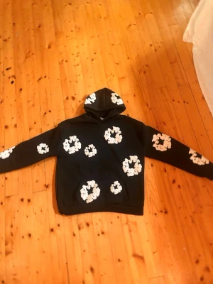 Svart hoodie Denim Tears - Säljer en svart hoodie från Denim Tears med stora vita blommor tryckta över hela tröjan, inklusive huvan och ärmarna. Hoodien har klassisk passform, stor ficka framtill och  Perfekt för dig som gillar streetwear och unik design.användit Max 3 gånger