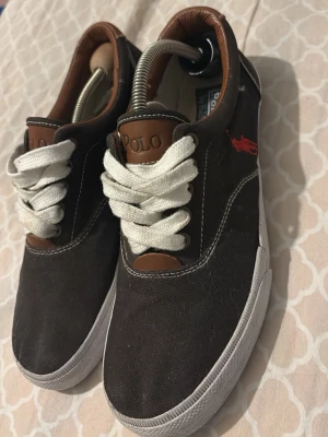 Ralph Lauren skor - Säljer ett par snygga sneakers från Polo Ralph Lauren i klassisk design. Skorna är i mörk färg med bruna detaljer och vit sula, vilket gör dem enkla att matcha till både vardag och lite mer uppklädda outfits. Den ikoniska röda loggan på sidan ger en exklusiv känsla.  ✔ Bekväma att gå i ✔ Klassisk och tidlös modell ✔ Fint skick (se bilder) ✔ Perfekta till både jeans och chinos