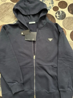 Mörkblå hoodie med dragkedja från Prada - Snygg mörkblå hoodie från Prada med dragkedja framtill och klassisk huva. Hoodien har en diskret Prada-logga på bröstet och praktiska fickor framtill. Tillverkad i mjuk bomull för en skön känsla. Perfekt för dig som gillar exklusiva streetwear-plagg.