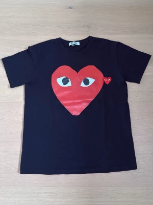 Svart Comme des Garçons Play t-shirt - Svart t-shirt från Comme des Garçons Play med det ikoniska röda hjärtat med ögon tryckt på bröstet och ett mindre hjärta bredvid. Klassisk passform och rund hals. Perfekt för dig som vill ha en statement tee med unik design.