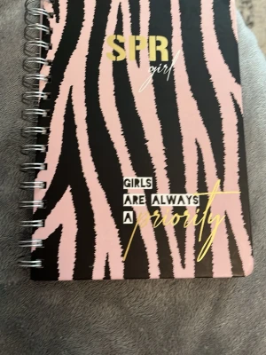 Spiralbunden SPR girl anteckningsbok - Cool anteckningsbok med spiralbindning och zebramönster i rosa och svart. Framsidan har texten 'Girls are always a priority' i guld och vitt. Perfekt för att skriva ner tankar, idéer eller planer. Omslaget är gjort av kartong.