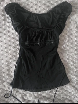 Svart snörad topp med spetsdetalj - Snygg svart topp med snörning i sidorna och spetsdetalj under bysten. Toppen har korta puffärmar och en figurnära passform som framhäver midjan. Passar storlek xs-s🩷