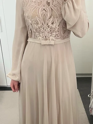 Lång ljusbeige klänning med spets - Elegant långklänning i ljusbeige med transparent spets upptill och broderade blommor. Klänningen har långa ärmar, ett tunt bälte med rosett i midjan och ett flytande kjolparti. Perfekt för speciella tillfällen där du vill känna dig extra fin.