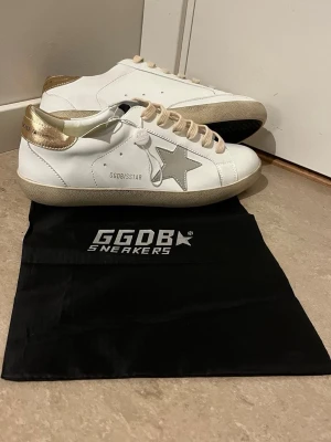 Golden Goose vita sneakers med guld - Golden Goose sneakers i vitt läder med metallicguld på hälen och beige sula. Klassisk låg modell med beige skosnören och grå stjärndetalj på sidan. Märkets logga syns på hälen och sidan. Snygga och trendiga skor som passar till det mesta.