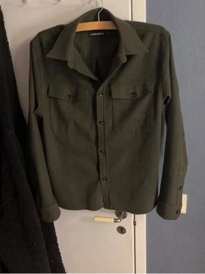 Mörkgrön overshirt från J.Lindeberg - Stilren mörkgrön overshirt från J.Lindeberg med klassisk krage, två bröstfickor med lock och knappar samt knäppning framtill. Skön och lite tjockare kvalitet som passar perfekt över en hoodie. Långärmad och enkel att matcha till olika outfits. Kan gå ner i pris vid snabb affär