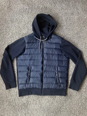 Marinblå Ralph lauren cardigan - Snygg marinblå hoodiejacka från Polo Ralph Lauren med quiltad front och två fickor med tryckknappar. Jackan har dragkedja, huva med dragsko och mjuka ärmar i sweatshirtmaterial. Perfekt för en avslappnad och sportig stil. Väldigt bra skick, bara en liten lagning som inte syns vid användning.