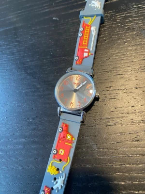 Grå klocka med brandbilsmotiv barn armbandsur - Cool klocka med grått armband i syntetmaterial och färgglada brandbilar som motiv. Urtavlan är silverfärgad med röda siffror och visare. Perfekt accessoar för dig som gillar lekfull och unik stil.