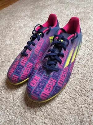 Adidas Messi lila/rosa inomhusfotbollsskor - Säljer ett par snygga Adidas Messi fotbollsskor i blått och rosa med gula detaljer. Skorna har texten 'Messi' tryckt över hela ovandelen, svarta snören och en neonrosa insida. Perfekta för dig som vill sticka ut på planen och gillar Messi. Storlek 40 2/3. Skriv vid intresse!