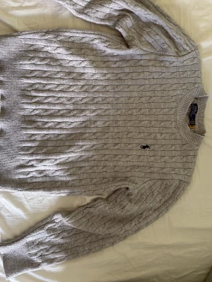 Ljusgrå kabelstickad tröja Polo Ralph Lauren - Kabelstickad tröja i ljusgrått från Polo Ralph Lauren med klassisk rund halsringning och broderad logga på bröstet. Tröjan har ribbade muddar vid ärmslut och nederkant. Perfekt för lager på lager och en clean look.