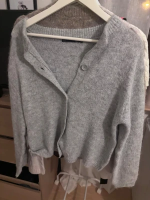 Ljusgrå stickad kofta med knappar - Mysig ljusgrå stickad kofta med  knappar framtill och ribbade muddar. Koftan har en avslappnad passform och är perfekt att slänga på sig när det är kyligt. Tillverkad i ett mjukt och värmande material.