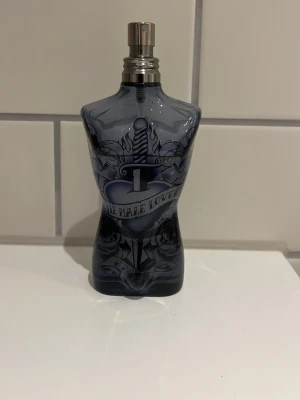 Jean Paul Gaultier Le Male Lover EdT - Unik parfymflaska formad som en manlig torso i mörkblått glas med tatueringsinspirerat motiv och metallisk spraykork. Doften är Le Male Lover Eau de Toilette, 125 ml. Flaskan har cool design och är perfekt för dig som gillar statement-parfymer.och de här lådan 