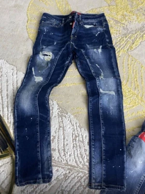  DSQUARDS Mörkblå slitna jeans med färgstänk - Säljer ett par mörkblå jeans med slitningar och färgstänk för en cool, trendig look. Jeansen har raka ben, klassisk femficksmodell och detaljerade slitningar på låren och knäna. Perfekta för dig som gillar streetstyle och vill sticka ut.