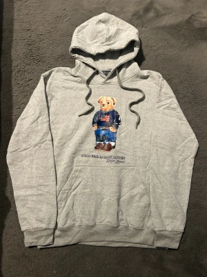 Grå hoodie Polo Bear Ralph Lauren - Snygg grå hoodie från Ralph Lauren med den ikoniska Polo Bear-trycket framtill. Hoodien har huva med dragsko och en stor magficka. Tillverkad i mjukt bomullsmaterial, perfekt för en avslappnad och trendig look.