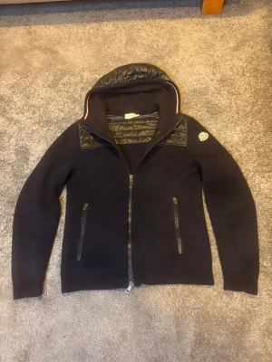 Mörkblå cardiganjacka från Moncler - Snygg mörkblå cardiganjacka från Moncler med quiltade detaljer på huva och axlar. Säljer den pågrund av att den har blivit förliten.  Jackan har dragkedja framtill, två fickor med dragkedja och Moncler-logga på ärmen. Materialmix av stickat tyg och glansigt nylon ger en modern look. Hör av dig vid minsta lilla fundering 