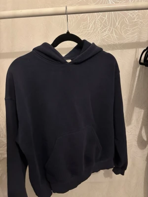 Mörkblå hoodie med magficka - En stilren mörkblå hoodie med huva och stor magficka framtill. Hoodien har en avslappnad passform och ribbade muddar vid ärmslut och nederkant. Perfekt för en chill och bekväm look.