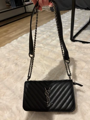 Svart YSL axelväska i läder - Säljer en fejk svart axelväska från YSL i quiltat läder med silverfärgad kedjerem och ikoniska YSL-loggan framtill. Väskan har en stilren, rektangulär form och dragkedja upptill. Perfekt för dig som vill ha en lyxig och trendig accessoar.