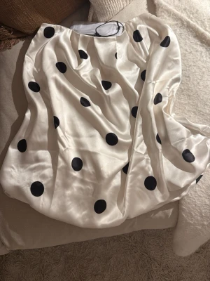 Vit prickig bandeau tubtopp - Snygg bandeau tubtopp i vit satin med stora svarta prickar. Toppens material är glansigt och mjukt, och den är axelbandslös med en loose passform. Perfekt för dig som vill ha en trendig och lekfull look. Aldrig använd med lapp på!