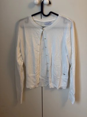 Vit kofta från Holly & White Lindex - Klassisk vit kofta från Holly & White med guldiga knappar framtill. Långärmad modell i tunt, mjukt material. Enkel och stilren design med liten logodetalj vid nederkanten. Koftan är knappt använd och i mycket bra skick.