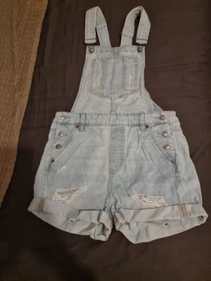 Ljusblå jeansshorts hängselshorts - Säljer ett par ljusblå jeansshorts med hängslen och slitna detaljer. Shortsen har upprullade benslut, bröstficka och flera knappar i sidorna. Perfekta för en avslappnad och trendig look.