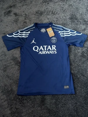 Blå PSG matchtröja Jordan x Qatar - Snygg blå PSG matchtröja i samarbete med Jordan och Qatar Airways. Tröjan har vita detaljer på axlarna, PSG-logga på bröstet och nummer 14 med D. Doué på ryggen. Tillverkad i lätt och funktionellt material, perfekt för fotboll eller träning.