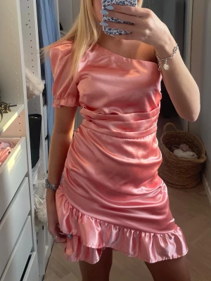 Rosa satin one shoulder klänning - Snygg kort rosa klänning i satin med volangkant nertill och en axel. Puffärm på ena sidan ger en trendig look. Perfekt för dig som vill sticka ut med en unik och feminin stil.