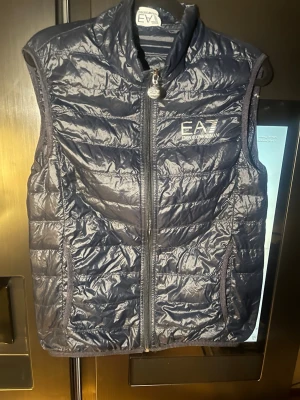 Svart dunväst från Emporio Armani EA7 - Snygg svart dunväst från Emporio Armani EA7 med quiltad design och dragkedja framtill. Västen har hög krage och två sidofickor. Perfekt för lager-på-lager och har en stilren logga på bröstet. Lätt och smidig att bära, passar till många outfits.