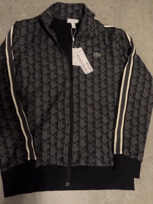 Lacoste monogram kofta - Svart track jacket från L@coste med hel dragkedja, vitt geometriskt mönster och vita ränder längs ärmarna. Klassisk ståkrage och broderad grön krokodil-logga på bröstet. Perfekt för en sportig och trendig look. Storlek L men sitter M