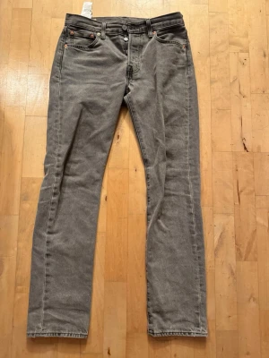 Grå 501 jeans Levi's - Säljer ett par grå jeans från Levi's med rak passform och klassisk femficksdesign. Jeansen har knappgylf, bälteshällor och detaljer i kopparfärg. Tillverkade i slitstark denim, perfekta för en avslappnad stil. Storlek 28/32