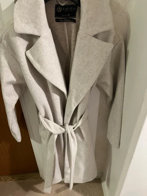 Ljusgrå kappa från Amisu med skärp - Stilren ljusgrå kappa från Amisu med breda slag och knytskärp i midjan. Kappan har en rak passform, två sidofickor och är tillverkad i ett mjukt tyg som ger en clean look. Perfekt för dig som gillar minimalistisk och trendig stil.