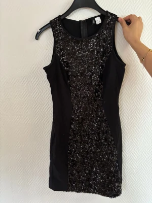 Svart paljettklänning från Divided - Säljer en svart kort paljettklänning från Divided. Klänningen är ärmlös och har glittriga paljetter framtill som ger en snygg partylook. Figurnära passform och rund halsringning. Perfekt för dig som vill sticka ut!