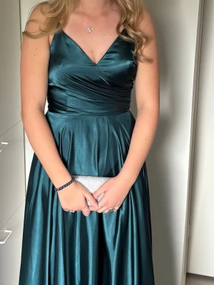 Mörkgrön långklänning med v-ringning - Elegant mörkgrön långklänning med v-ringad omlott-topp och smala axelband. Klänningen har en glansig finish och markerad midja som ger en snygg siluett. Perfekt för bal eller festligare tillfällen. Klänningen är från Ivoryfield och har ett ny pris på 3300. Använd endast en gång och är kemtvättad. Jag är 175 cm och den är hela vägen ner till marken. Säljer även skor och väska till i en annan annons. Går bra att hämta upp hos mig. 