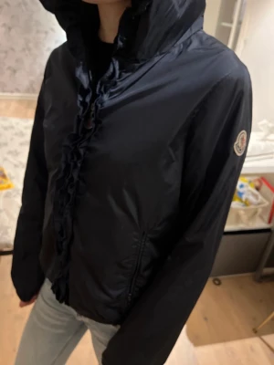 Mörkblå vindjacka från Moncler - Snygg mörkblå vindjacka från Moncler med huva och dragkedja framtill. Jackan har Moncler-logga på ärmen och diskreta fickor med dragkedja. Tillverkad i lätt, glansigt material som ger en sportig look. Perfekt för blåsiga dagar.