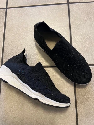 Svarta slip-on sneakers med blommor - Säljer ett par svarta slip-on sneakers med stickad ovandel och dekorativa blommor i svart. Skorna har vit sula och elastisk mudd runt ankeln för enkel påtagning. Perfekta för dig som gillar en sportig men ändå stilren look.