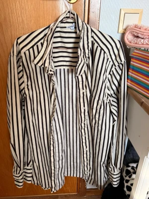 Randig skjorta från Sincerely Love - Säljer en vit skjorta med svarta vertikala ränder från Sincerely Love. Skjortan har klassisk krage, långa ärmar och knäppning framtill. Perfekt för dig som gillar en stilren och trendig look.