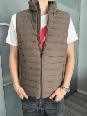 Brun dunväst från Jack & Jones - Snygg brun dunväst med horisontella sömmar och hög krage. Västen har dragkedja framtill och två sidofickor. Storlek s, men passar som m. Enkel och stilren design utan synligt märke.