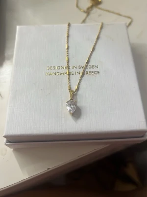 Guldhalsband med hjärtformad sten Syster P - Elegant halsband i guld med en gnistrande hjärtformad klar sten som hänge. Kedjan är tunn och har små detaljer längs länkarna. Designad i Sverige och handgjord i Grekland. Perfekt för dig som gillar stilrena och romantiska smycken. Köptes förra månaden för 1395 kr men säljer eftersom jag är mer av en silver tjej
