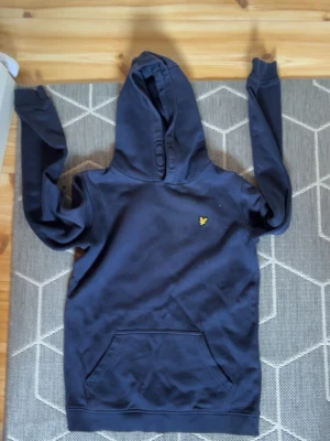 Mörkblå hoodie från Lyle & Scott - Snygg mörkblå hoodie från Lyle & Scott med klassisk gul logga på bröstet. Hoodien har stor huva och en praktisk magficka framtill. Tillverkad i mjukt bomullsmaterial och har ribbade muddar vid ärmslut och nederkant. strl 12-13 år men passar allt från 158-170