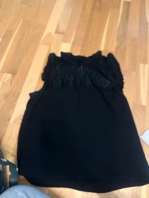 Svart spetsdetaljerad blus - Snygg svart blus med spetsdetaljer upptill och volang vid axlarna. Hög hals med liten öppning framtill ger en elegant touch. 