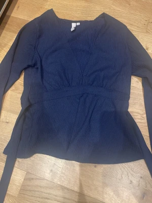 Mörkblå Nelly blus💙 - Säljer en jätte fin mörkblå blus med djup v-ringning och långa ärmar. Blusen har knytband i midjan och är tillverkad i ett lätt, strukturerat tyg som ger en trendig look. Har använt blusen cirka 5 gånger. Blusen är från Nelly och är trendig på Tik Tok. 😊💜