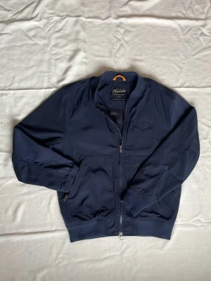 Marinblå bomberjacka Morris - Riktigt snygg marinblå bomberjacka från Morris med ribbade muddar, dragkedja framtill och två fickor med dragkedja. Jackan har en klassisk bomberkrage och diskret Morris-logga på bröstet. Materialet är slätt och lätt, perfekt för vår och höst. Hör av dig vid funderingar⁉️