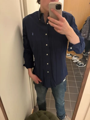 Mörkblå skjorta från Ralph Lauren - Klassisk mörkblå skjorta från Ralph Lauren med button-down krage och broderad logga på bröstet. Skjortan har långa ärmar och vita knappar framtill. Perfekt till jeans eller chinos för en stilren look.