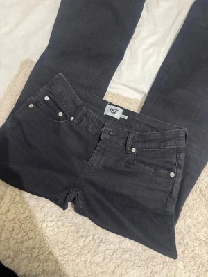 Svarta bootcut jeans från 157 - Svarta jeans från 157 med klassisk femficksmodell och bootcut-ben. Jeansen har silverfärgade nitar och knapp, samt bälteshällor i midjan. Perfekta för dig som gillar en tidlös och enkel stil.