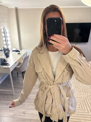 Beige trenchcoat med knytskärp - Snygg beige trenchcoat med klassisk krage och knytskärp i midjan. Jackan har några mörka fläckar på ryggen