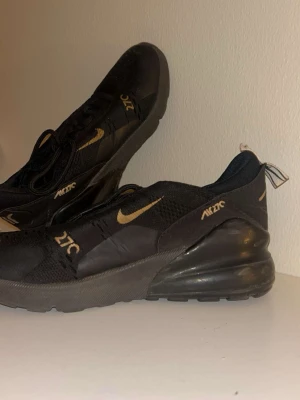 Nike Air Max 270 svarta/guld sneakers - Nike Air Max 270 sneakers i svart med guldiga detaljer och loggor. Skorna har en luftig mesh-överdel, synlig Air Max-dämpning i hälen och klassisk snörning. Snygg och sportig design som sticker ut med sina guldaccenter.