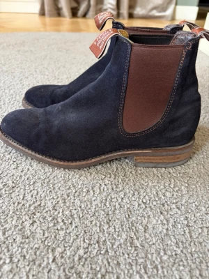 Svarta Chelsea boots i mocka - Säljer ett par klassiska Chelsea boots från R.M. Williams i svart mocka med bruna elastiska paneler och dragflikar med logga. Skorna har låg klack och rundad tå, samt en sula i brunt läder. Perfekta för en stilren look. Nypris 5000kr!!! 