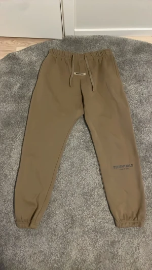 Beige mjukisbyxor från Essentials - Säljer ett par snygga beige mjukisbyxor från Essentials med dragsko i midjan och resår vid bensluten.STORLEK ÄR INTE RÄTT. HAR BLIVIT MINDRE ÖVER TID SÅ DEN KAN PASSA FRÅN M TILL L. Byxorna har en clean look med diskret logga på benet och är tillverkade i mjukt bomullsmaterial. Perfekta för chill dagar eller streetwear-stil.