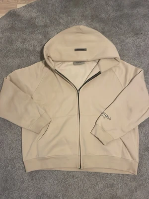 Beige Essentials hoodie med dragkedja - Säljer en beige hoodie från Essentials med stor huva och svart logotyp på ryggen. Hoodien har dragkedja framtill, ribbade muddar och fickor på sidorna. Materialet är mjukt och bekvämt, perfekt för en avslappnad look. Priset kan diskuteras.