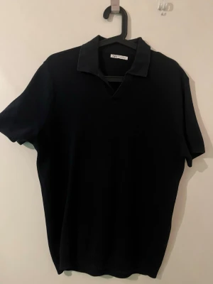 Svart polotopp från Zara Man - Stilren svart polotopp från Zara Man med krage och v-ringning. Toppen har korta ärmar och är tillverkad i ett mjukt, stickat material som ger en snygg och bekväm passform.