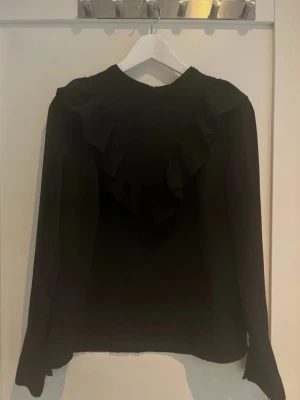 Svart blus med volangdetalj - Snygg svart blus med lång ärm och volangdetalj över bröstet och axlarna. Blusen har en rak passform och rund halsringning. Perfekt för dig som gillar stilrena plagg med en twist.