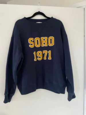 Mörkblå sweatshirt SOHO 1971 - Mörkblå sweatshirt med gul text 'SOHO 1971' på bröstet.